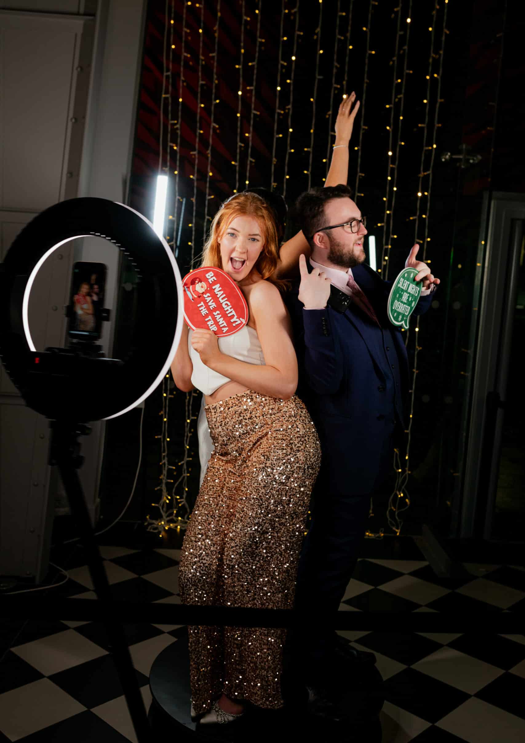 Man and woman posing playfully in a photo booth, holding fun props with festive lighting and a ring light in the background, showcasing a vibrant photo booth experience for events.