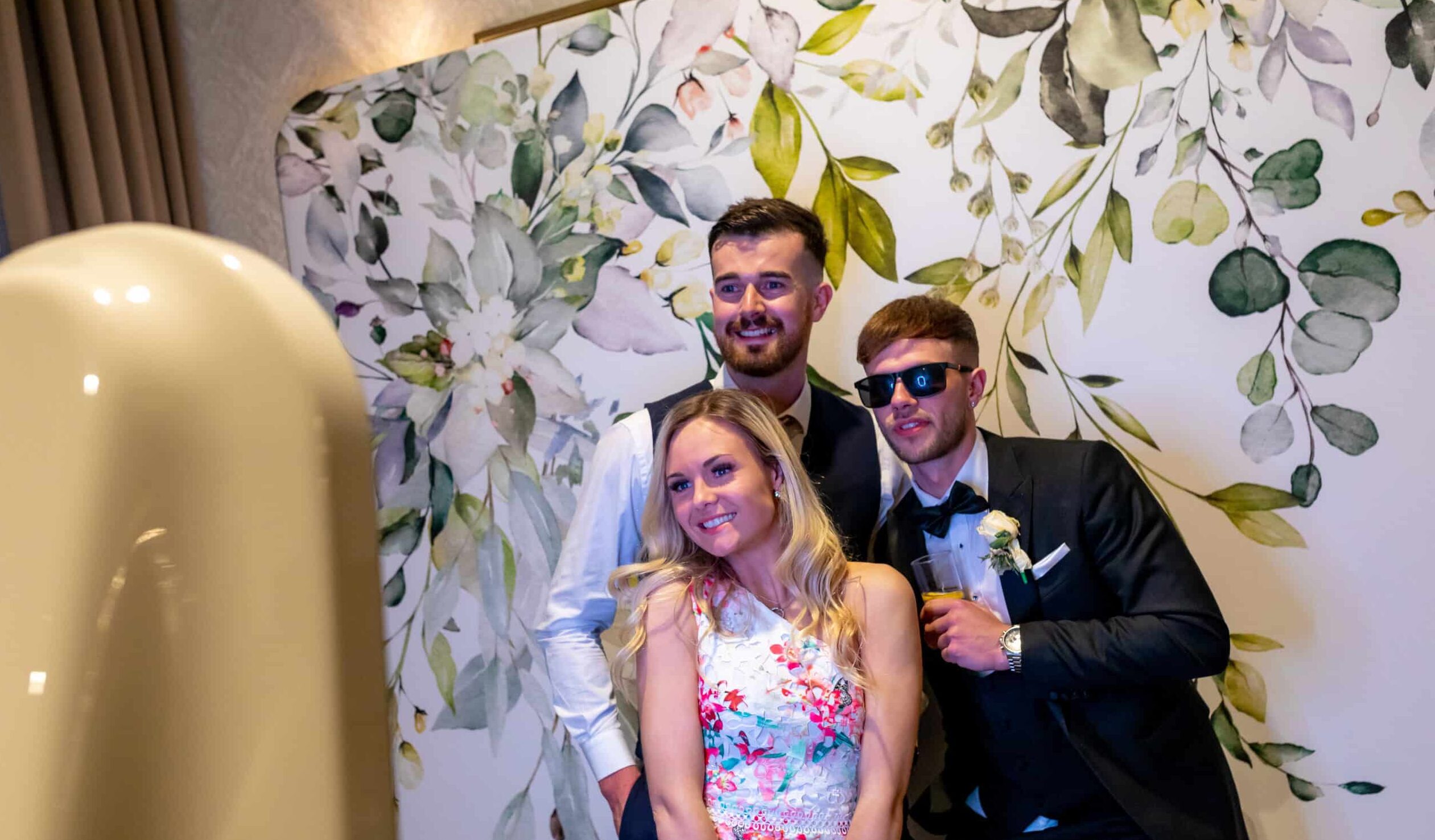 A woman in a formal dress posing in front of a beautiful flower wall at a wedding or corporate event.