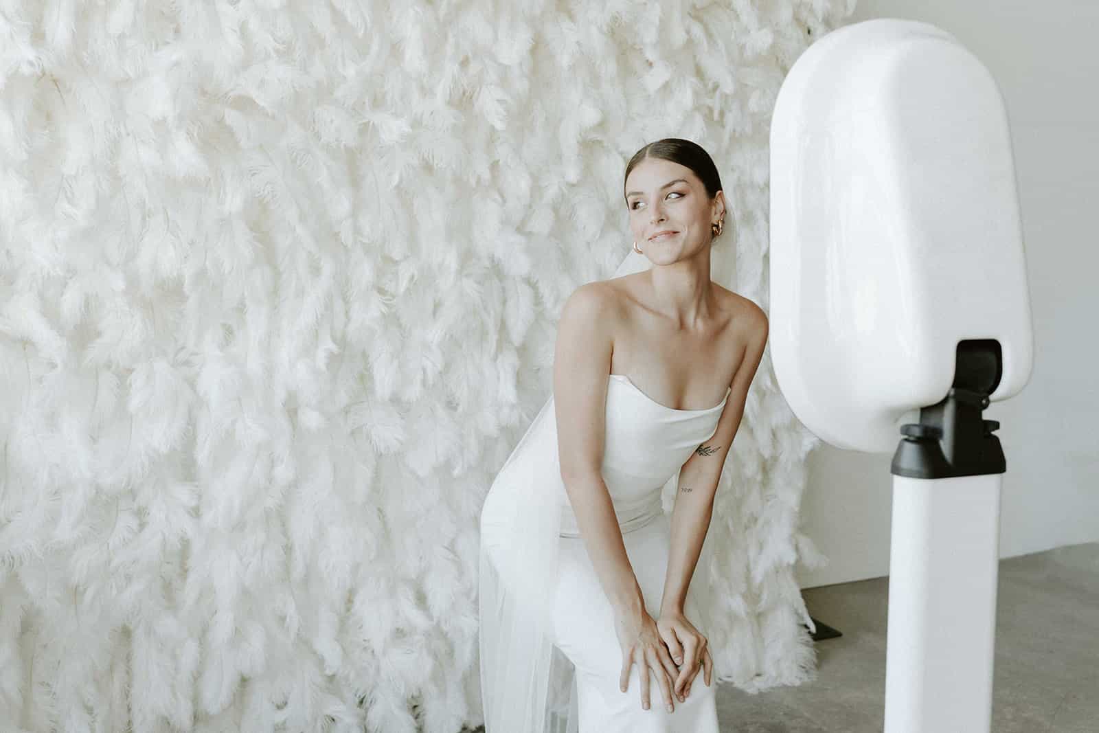 A couple smiling and posing for a picture at their wedding, with a white feather wall and the photo booth's lighting rig visible.