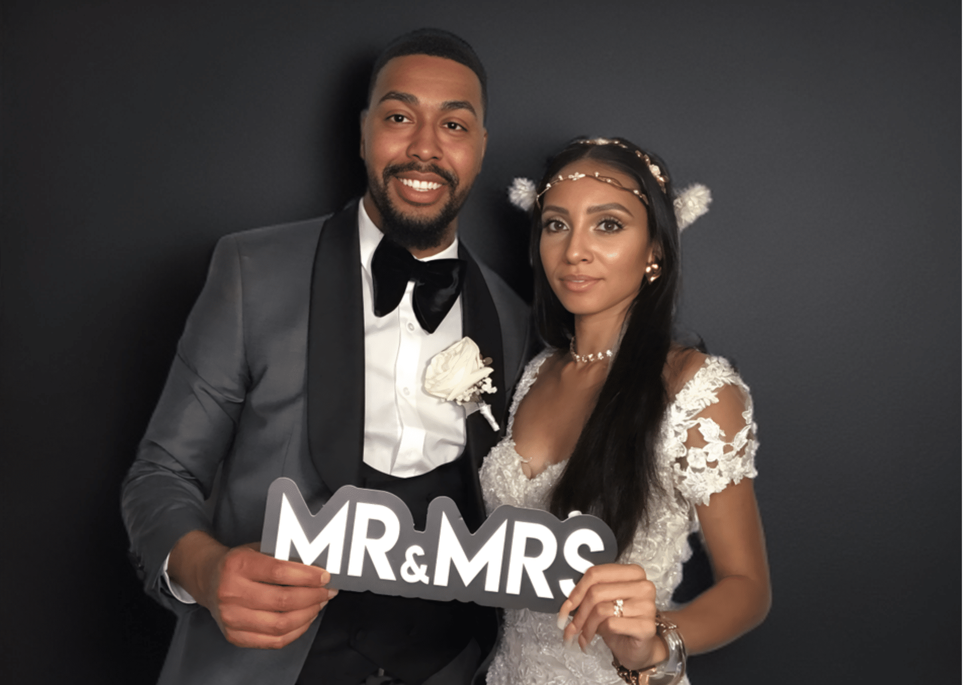 A bride and groom pose together in front of a black backdrop. The groom is wearing a gray tuxedo jacket with a black lapel, black bow tie, and white boutonniere. The bride is in a lace wedding gown with cap sleeves and a jeweled headband. They are holding a “MR & MRS” sign and smiling at the camera.