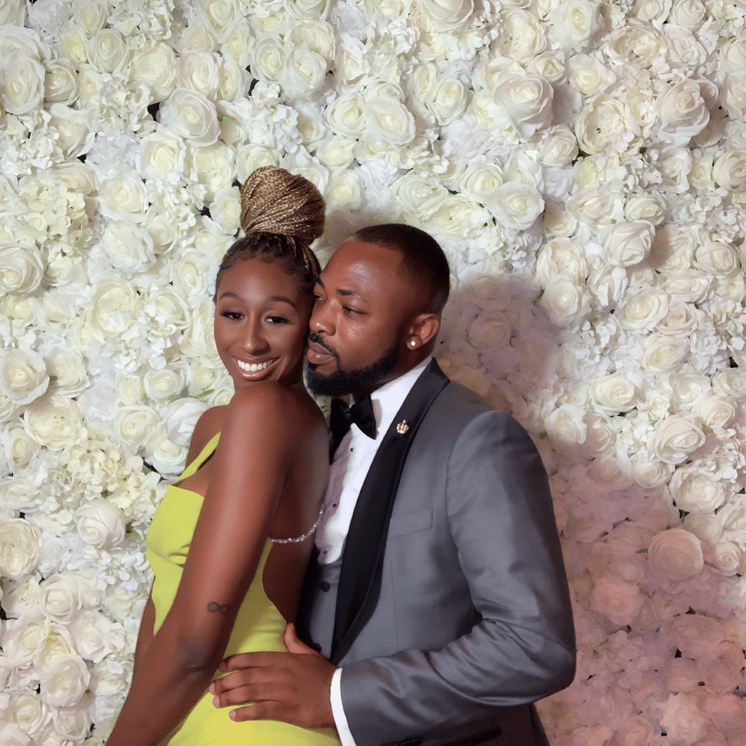 Couple posing in front of a floral backdrop, showcasing elegant attire; woman in a bright yellow dress and man in a gray tuxedo, capturing a joyful moment at a celebration.