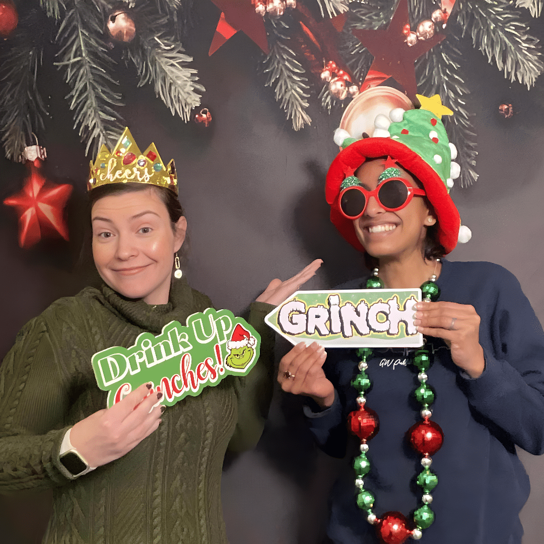 Two women posing with festive props, including "Drink Up Grinches!" and "GRINCH" signs, wearing holiday-themed accessories like a crown and a Christmas tree hat, against a backdrop of Christmas decorations, perfect for creating Instagram-worthy moments at events.