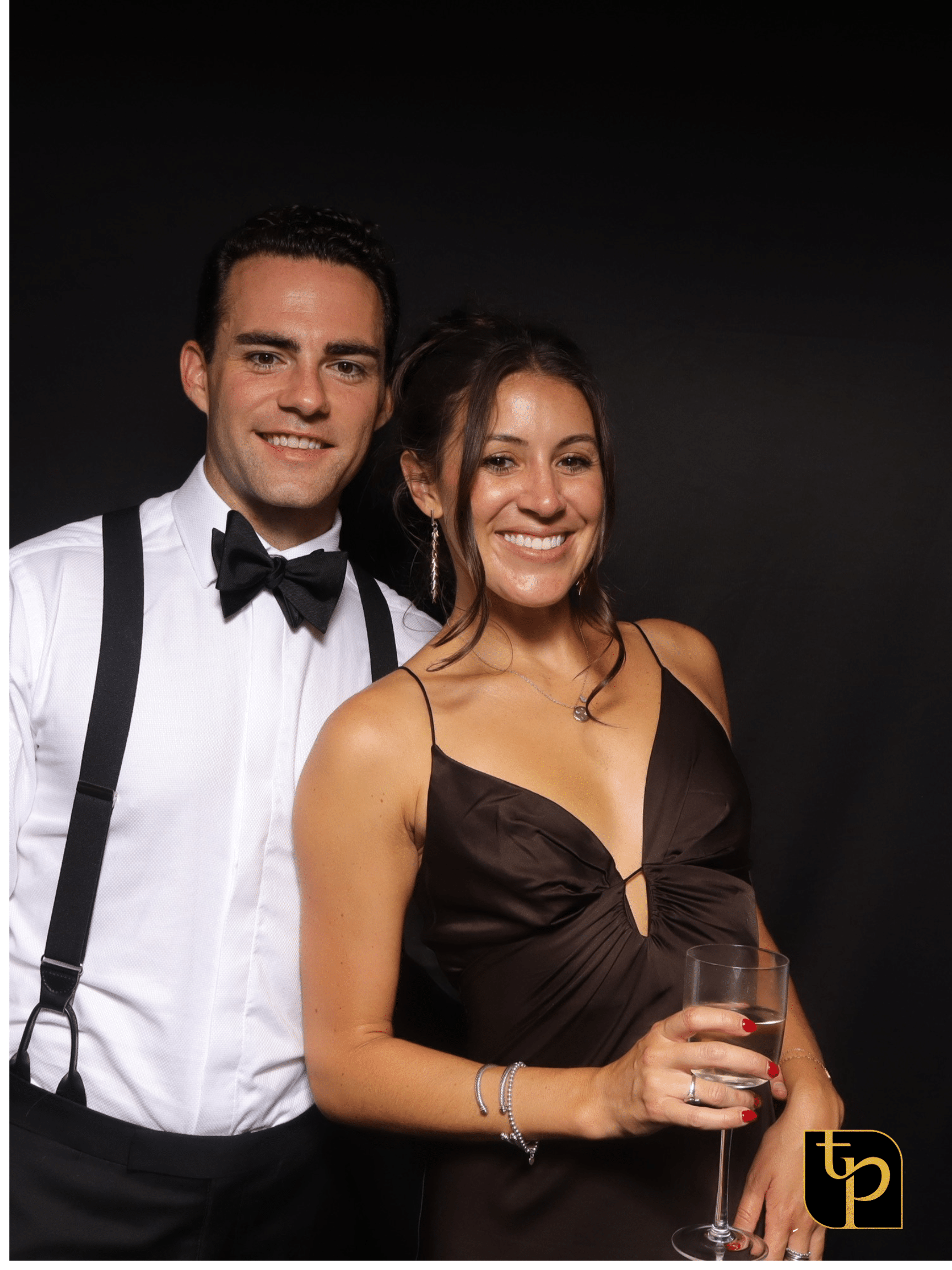 Couple posing in formal attire at a photo booth, showcasing a fun and engaging event experience, with a branded logo in the corner.