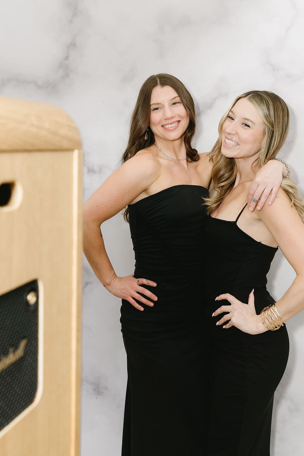 Two women in black dresses posing joyfully in front of a photo booth at an event, showcasing engagement and fun in a conference setting.