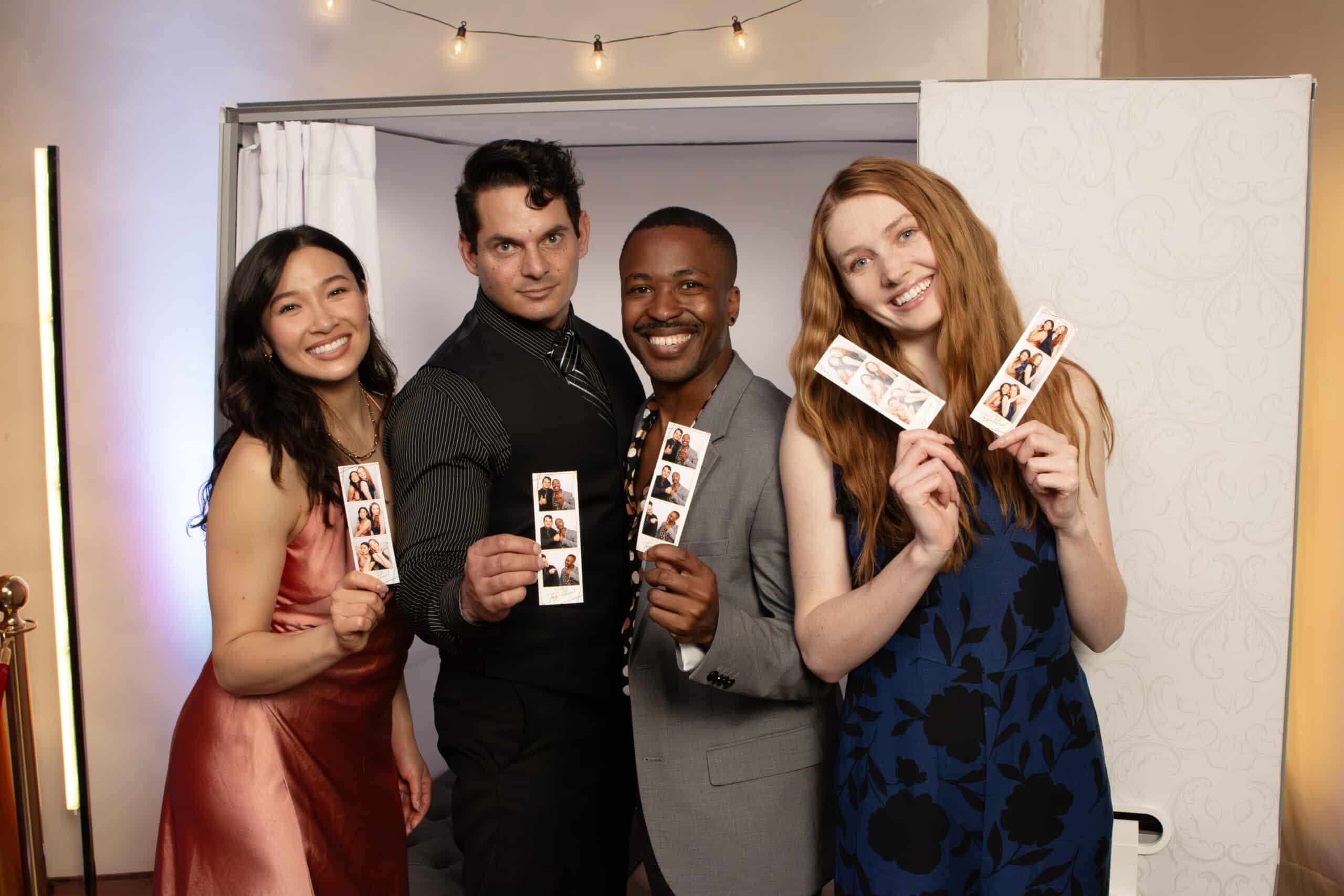 Group of four smiling individuals holding photo booth strips, showcasing fun keepsakes from an event, with a modern photo booth in the background, highlighting the social sharing aspect of The Phototique's services.