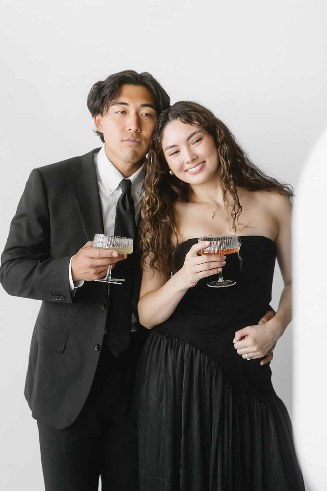 Couple holding champagne glasses at a photo booth, dressed in formal attire, smiling, with a simple white backdrop, enhancing event engagement and social sharing.