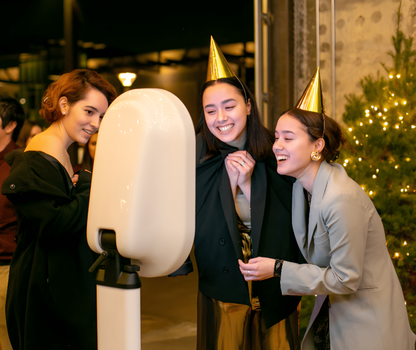 Three young women in party hats enjoying a photo booth experience, smiling and posing in a festive setting with soft lighting and holiday decorations.