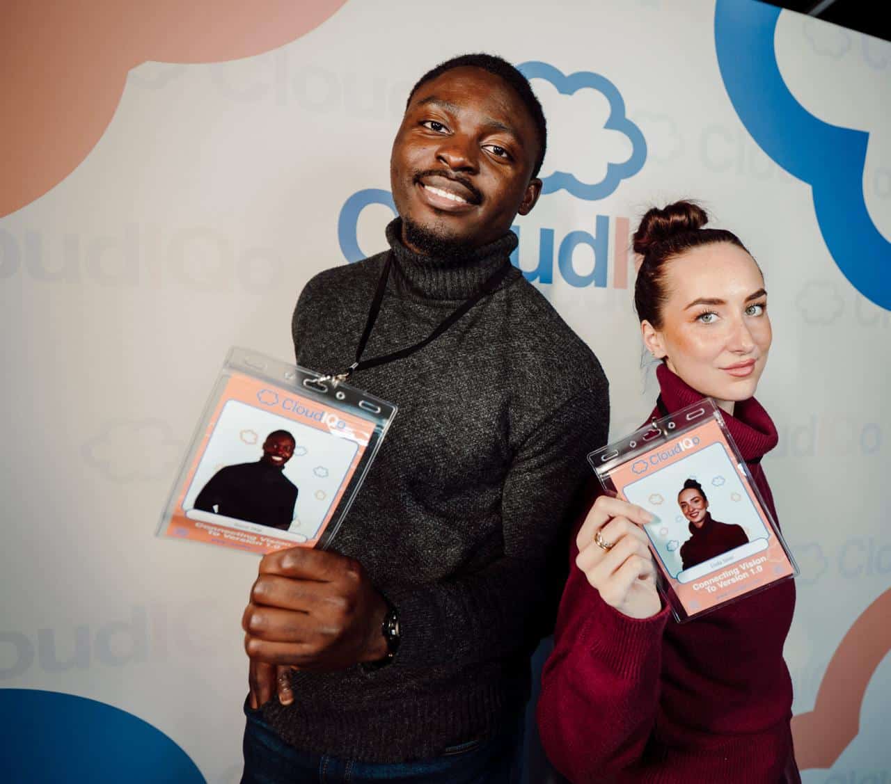 Two smiling attendees holding convention badges in front of a CloudQo branded backdrop, showcasing their identification for networking at an event.