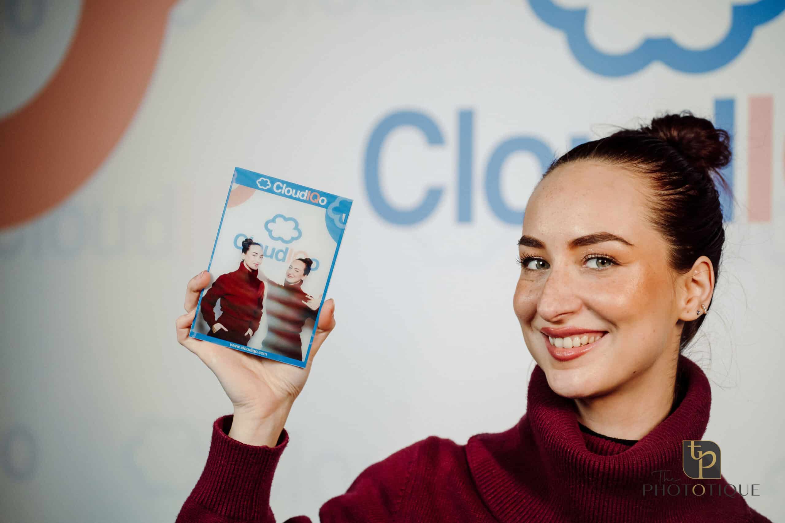 Woman smiling and holding a photo booth print featuring two attendees, with a branded backdrop displaying "CloudIQ," illustrating attendee engagement at a conference or trade show.
