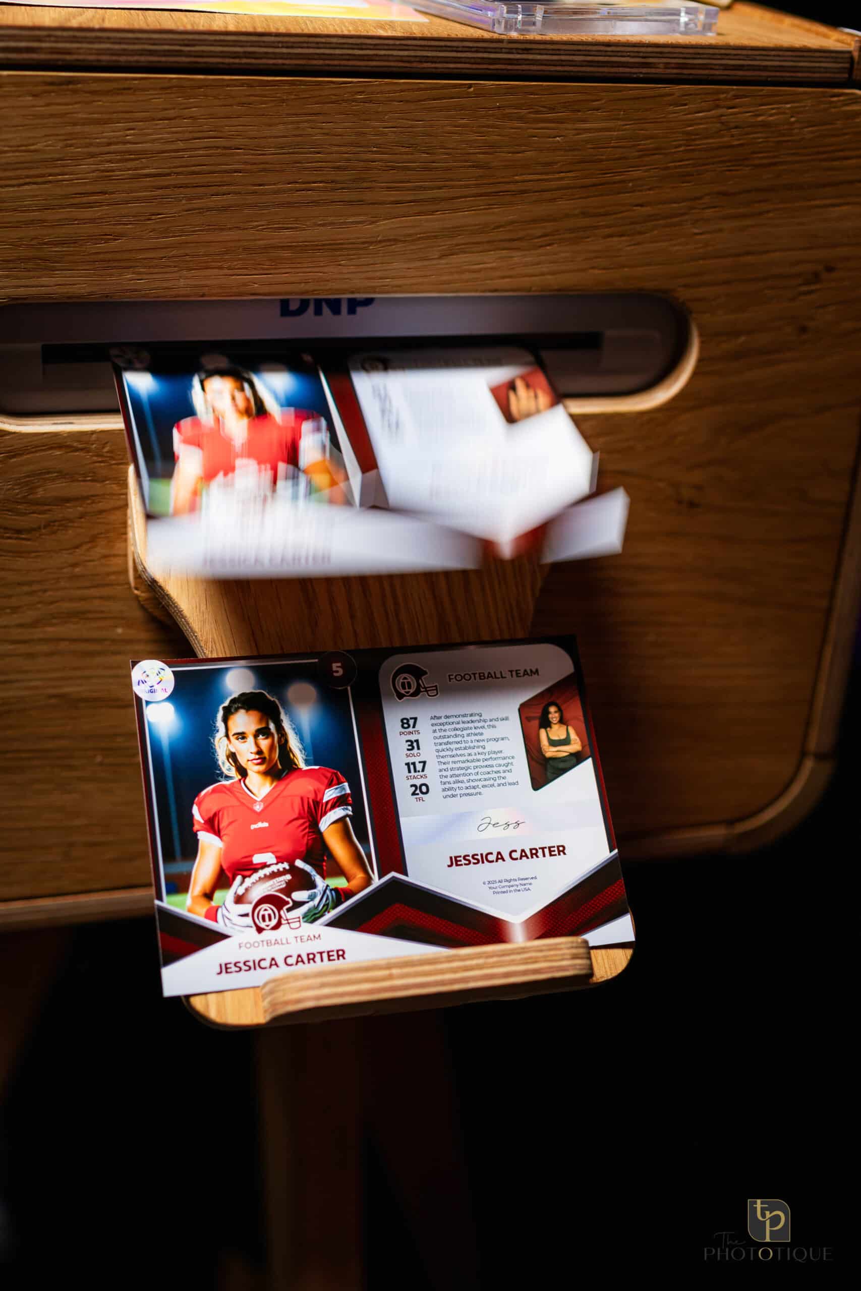 Trading cards featuring a female athlete in a red football uniform, displaying stats and personal information, emerging from a photo booth, emphasizing event experiences by The Phototique.