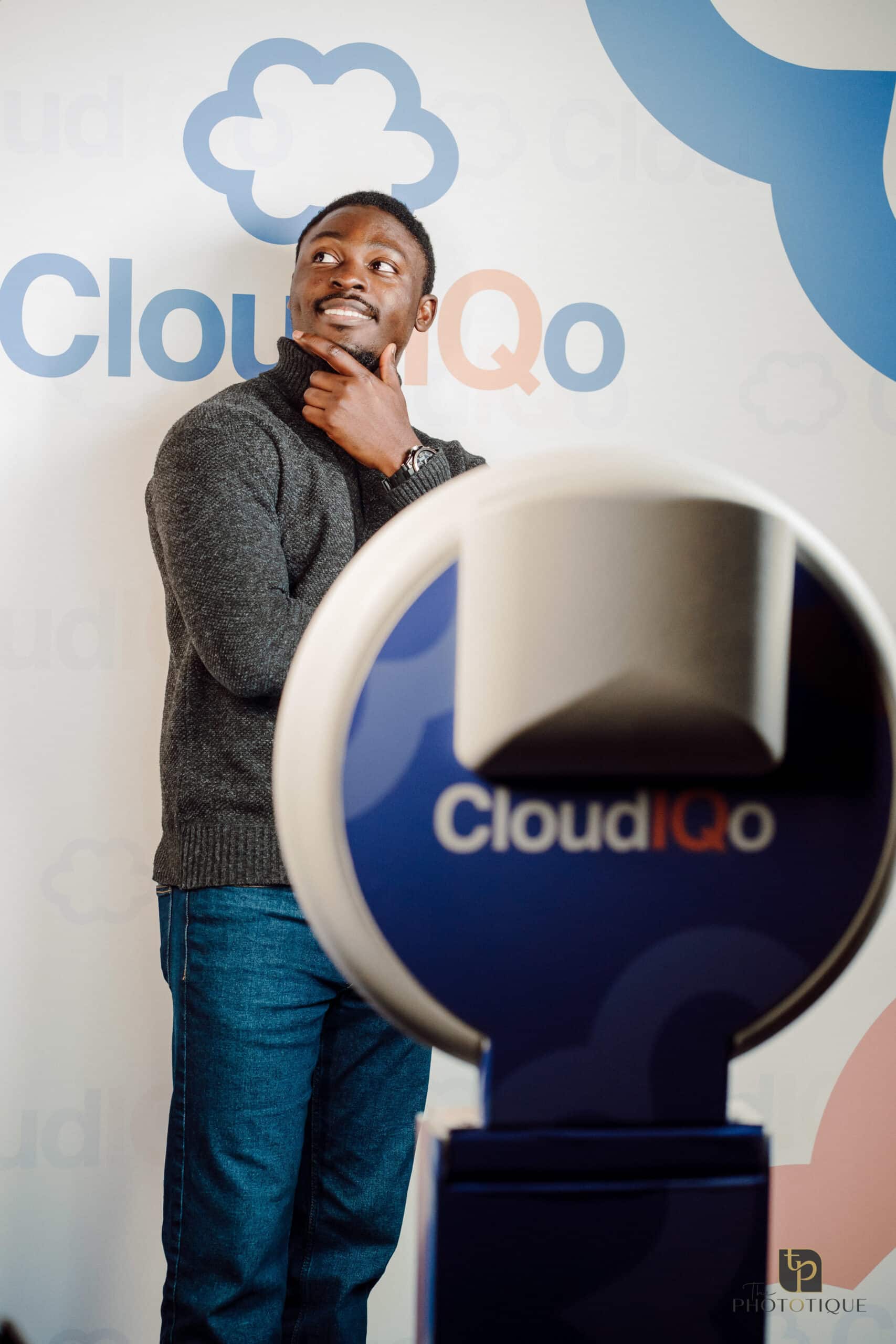 Man posing thoughtfully next to a photo booth branded with "CloudIQo," against a backdrop featuring cloud graphics, emphasizing event experiences and photo booth services.