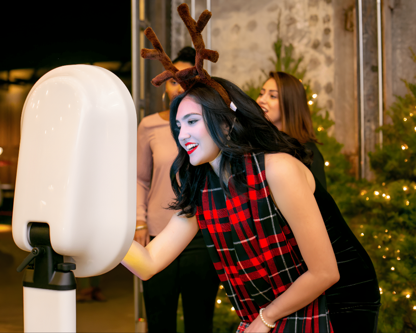 Woman in festive plaid scarf and reindeer antlers engaging with a holiday photo booth, surrounded by Christmas trees and party guests, capturing joyful moments at a seasonal celebration.