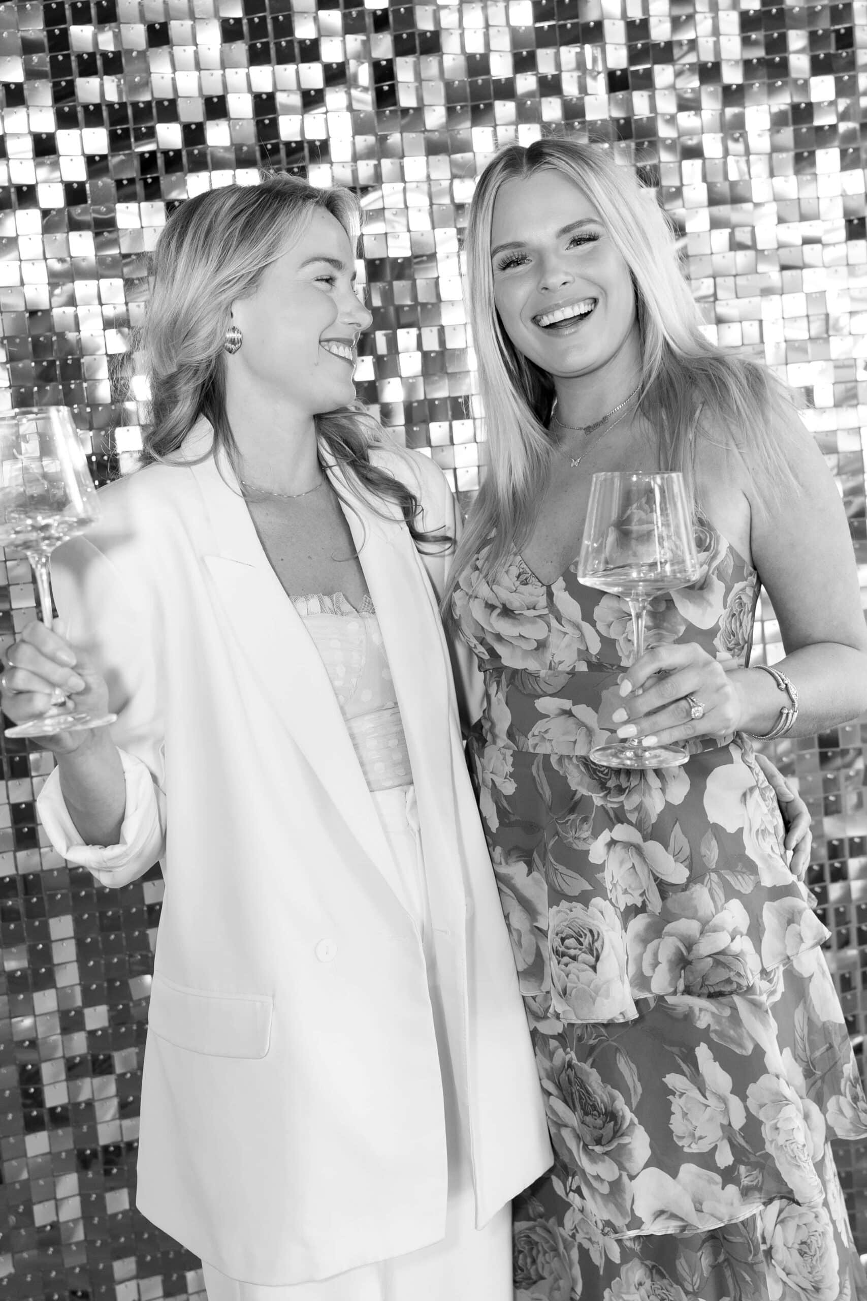 Two women smiling and holding wine glasses in front of a shimmering photo booth backdrop, ideal for events like weddings and celebrations.
