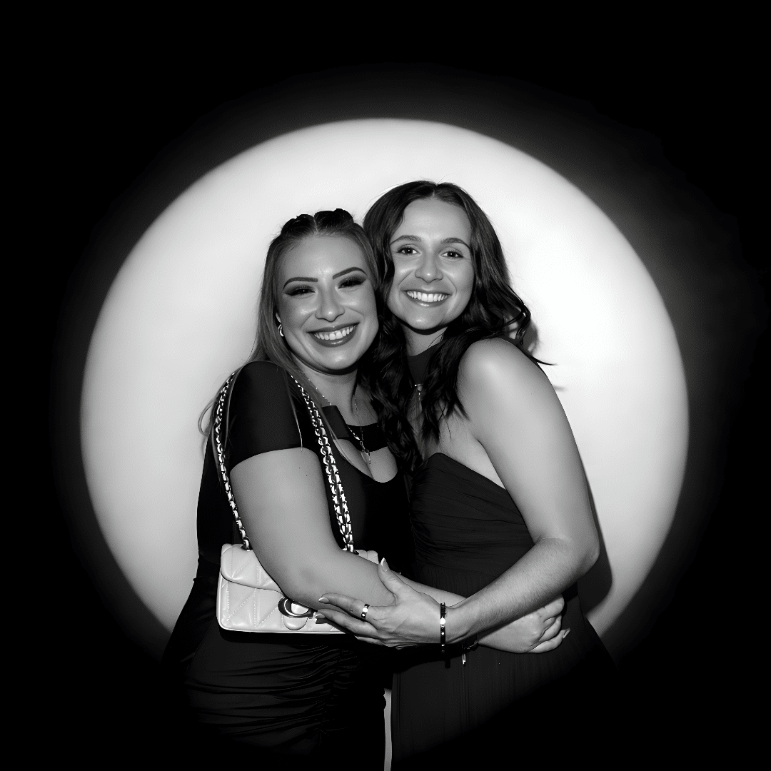 Two women smiling and embracing in front of a dramatic spotlight, showcasing the editorial-style photography of the Spotlight Booth experience by The Phototique.