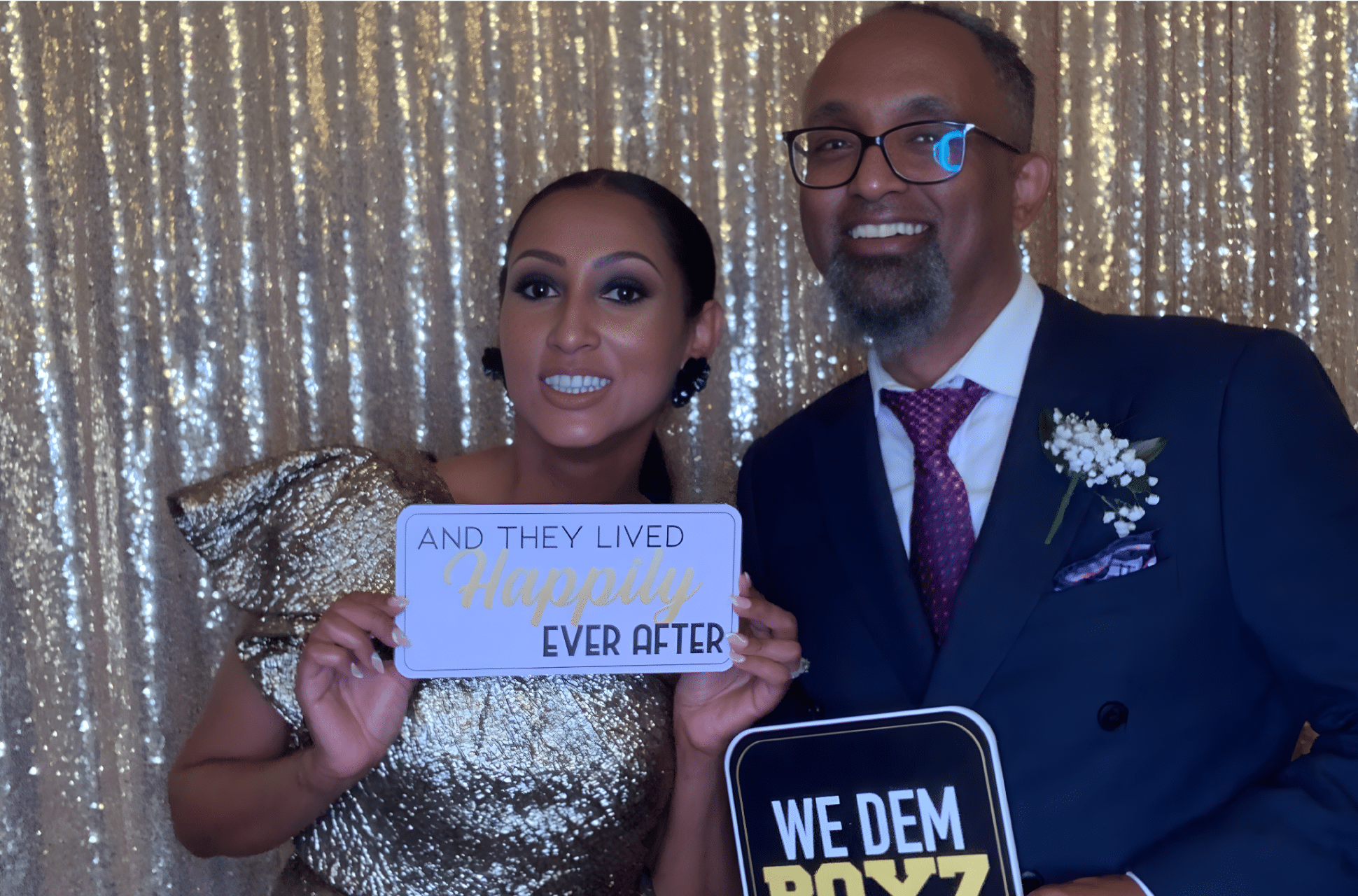 Couple posing in front of a shimmering backdrop, holding signs that read "AND THEY LIVED Happily Ever After" and "WE DEM BOYZ," showcasing a joyful moment at a wedding or event, reflecting The Phototique's customized photo booth experience.
