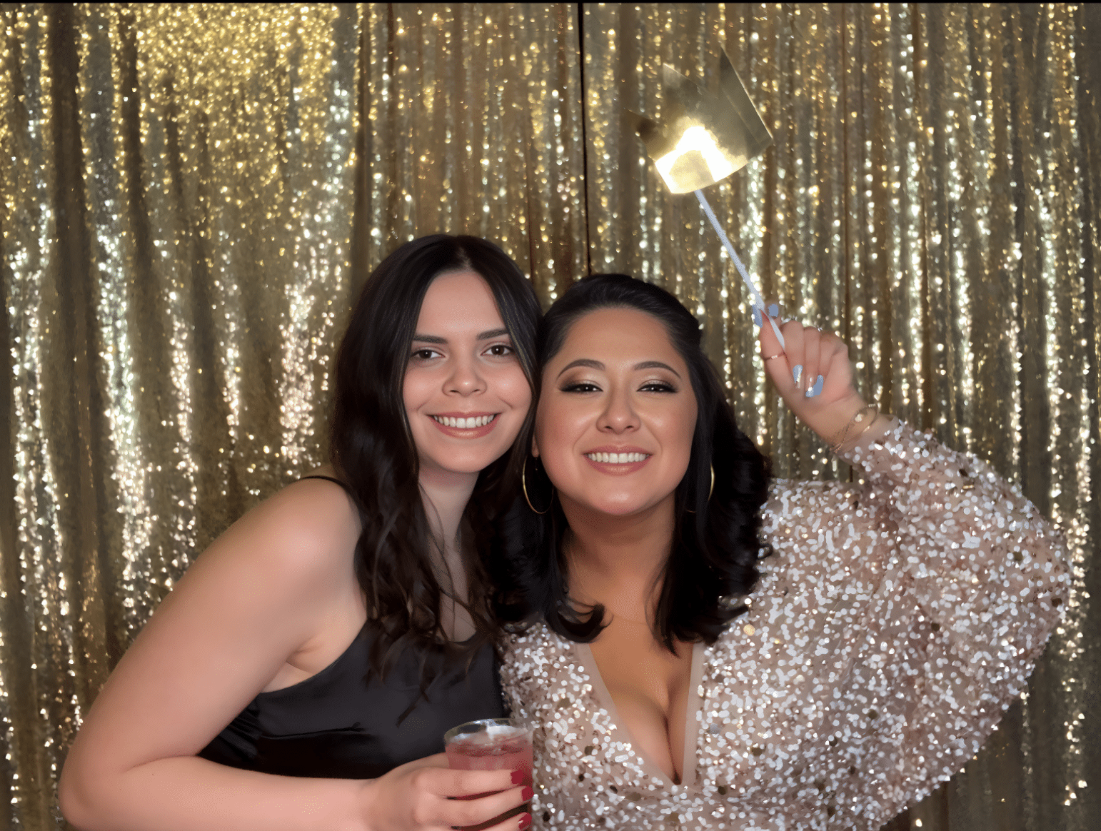 Two women posing joyfully in a photo booth, one holding a drink and the other a gold crown prop, with a sparkling gold backdrop enhancing the festive atmosphere, reflecting The Phototique's innovative event experiences in Washington, DC.