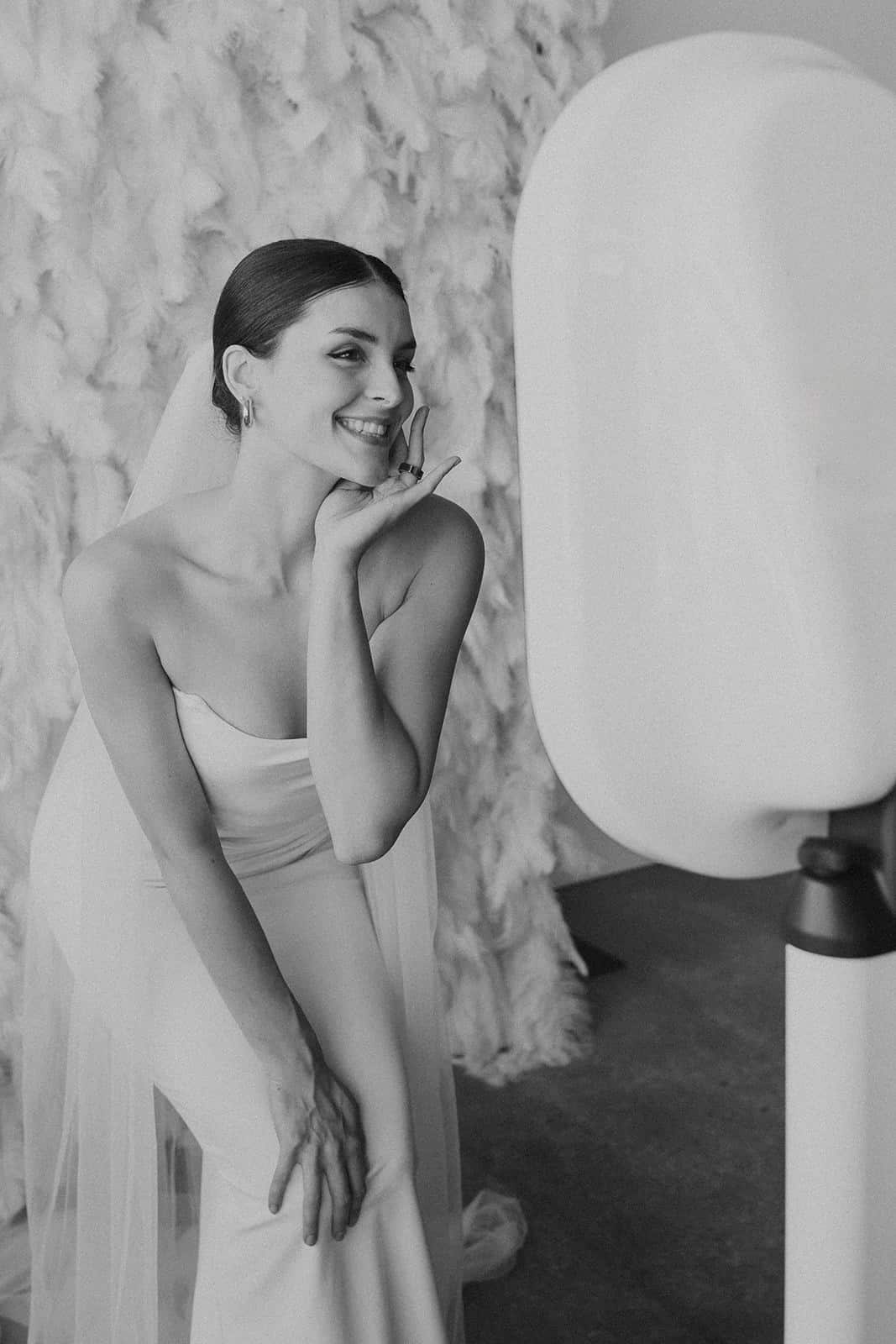 Bride posing playfully in front of a photo booth mirror, styled backdrop of feathers, showcasing elegant wedding portrait experience.