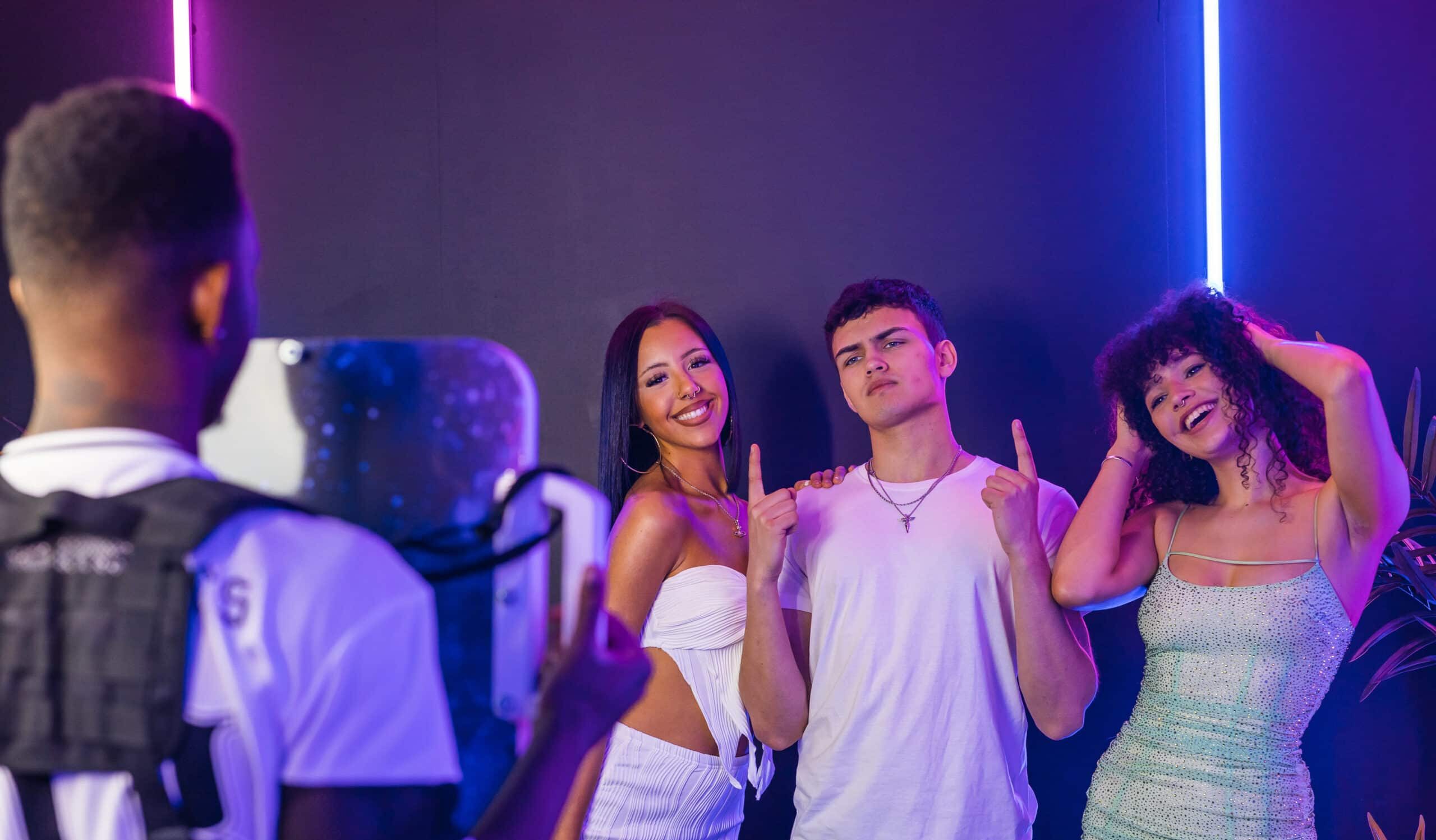 Group of three young adults posing playfully in front of a photo booth with vibrant purple and blue lighting, showcasing a fun event atmosphere.