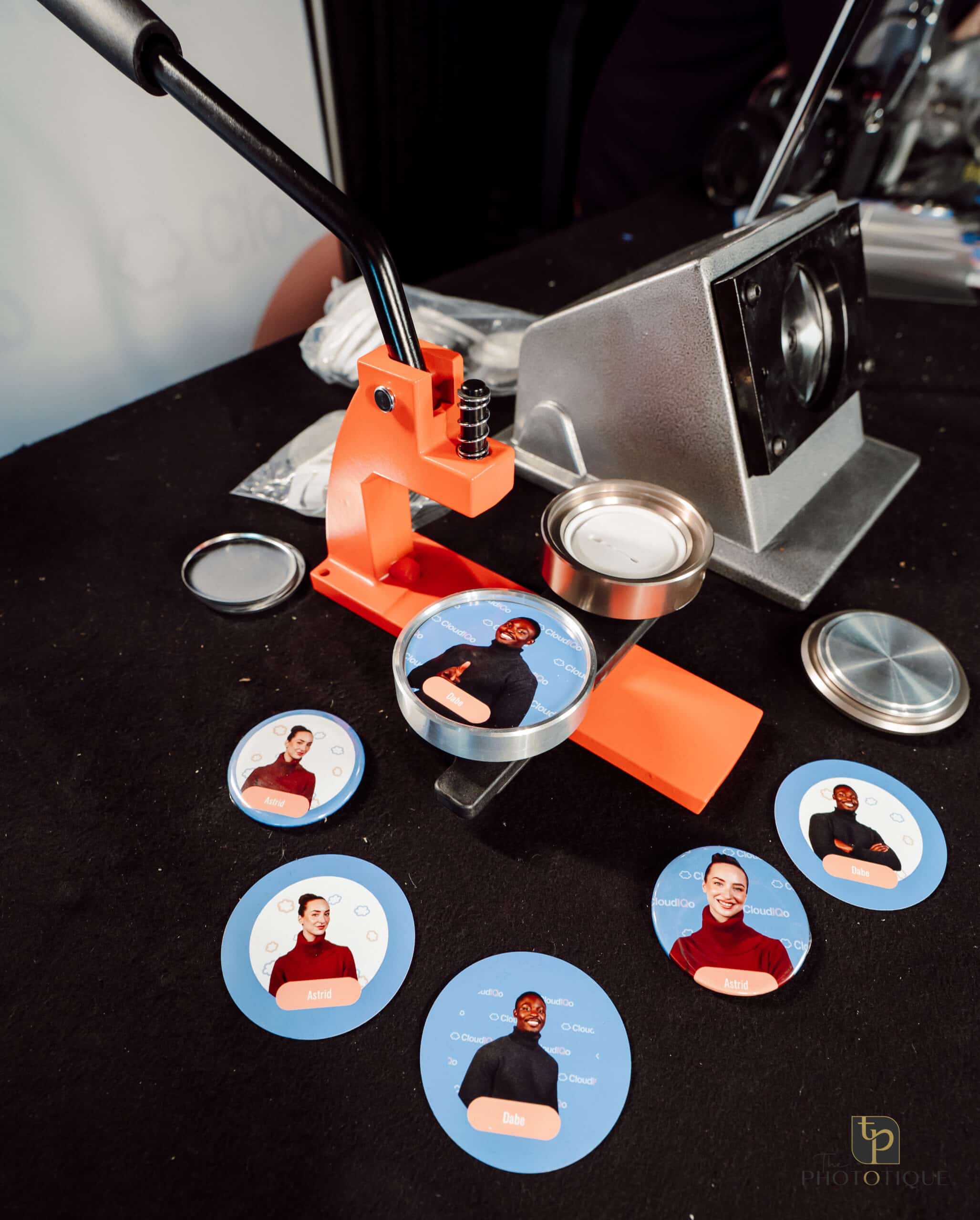 Merch booth equipment for creating custom keepsake buttons, featuring an orange button press and displayed buttons with images of guests, emphasizing interactive brand experiences.