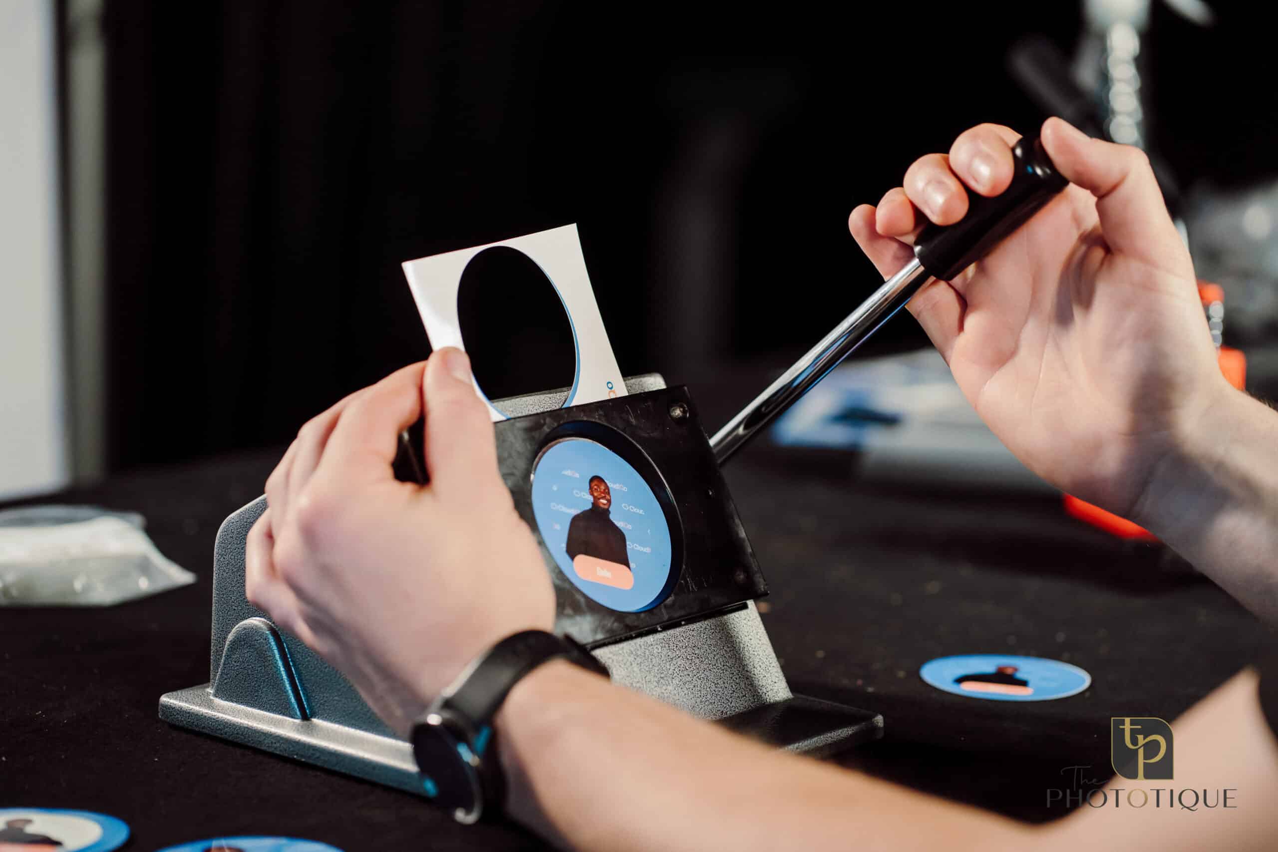 Hands operating a badge-making machine, creating custom keepsake merchandise for events, featuring a circular badge with a blue background and a photo.