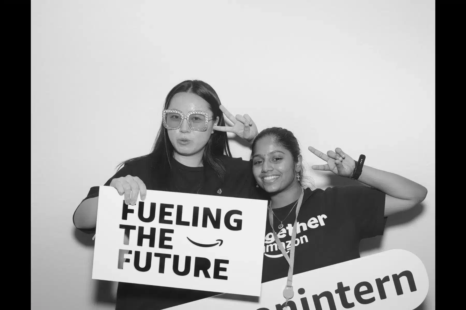 Two young women posing in a photo booth, one holding a sign that reads "FUELING THE FUTURE" and the other displaying a "together Amazon intern" sign, both smiling and making peace signs, reflecting a fun and engaging event atmosphere.