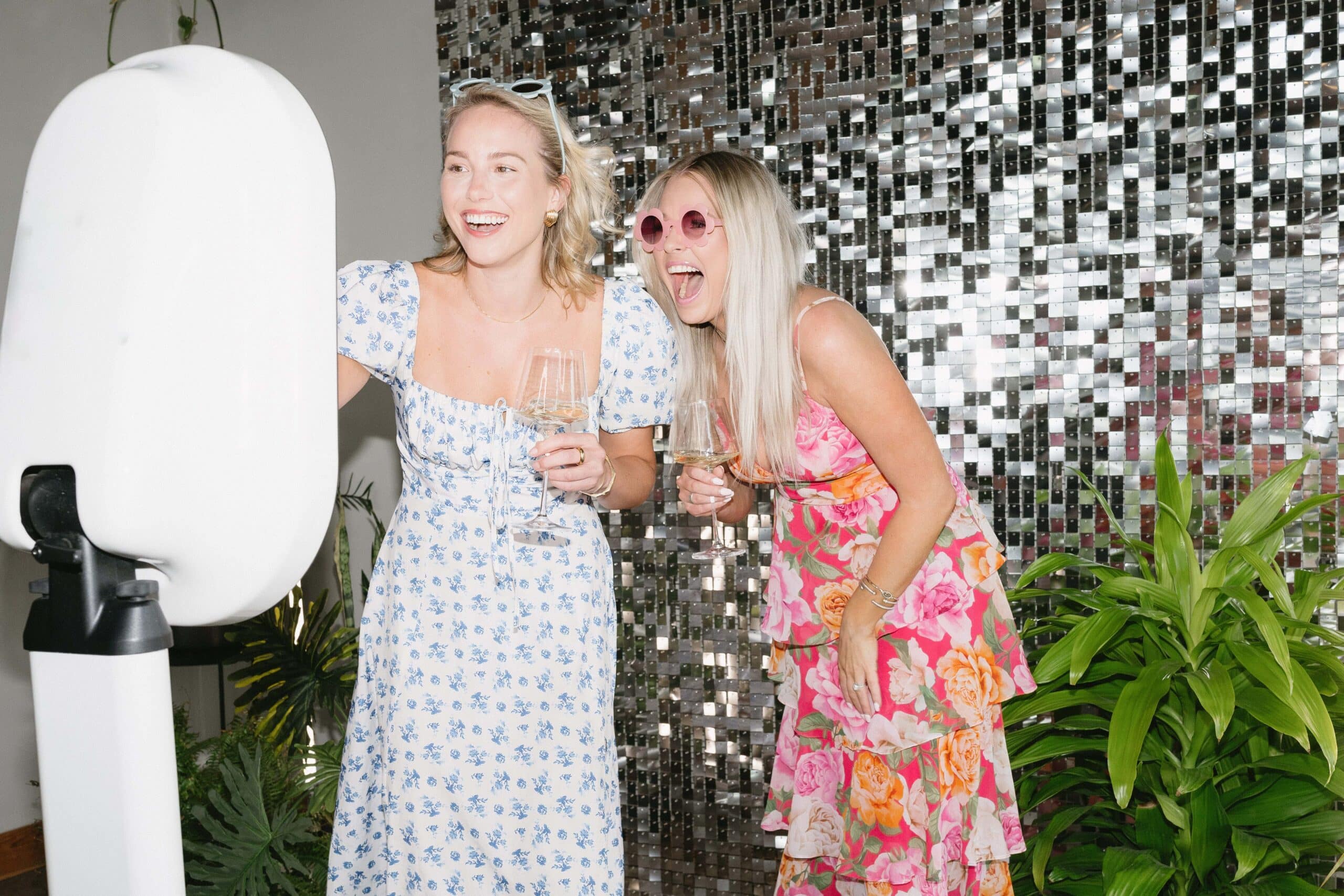 Two women enjoying a photo booth experience, smiling and holding glasses of champagne, with a sparkling mirrored backdrop and greenery, highlighting The Phototique's immersive event services.