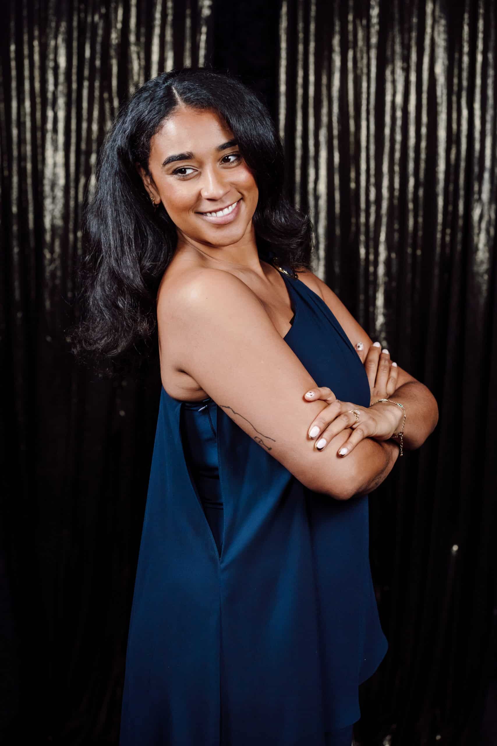 Woman smiling in a navy dress with arms crossed, posing against a shimmering black backdrop, capturing a glamorous moment suitable for red carpet events and high-profile celebrations.