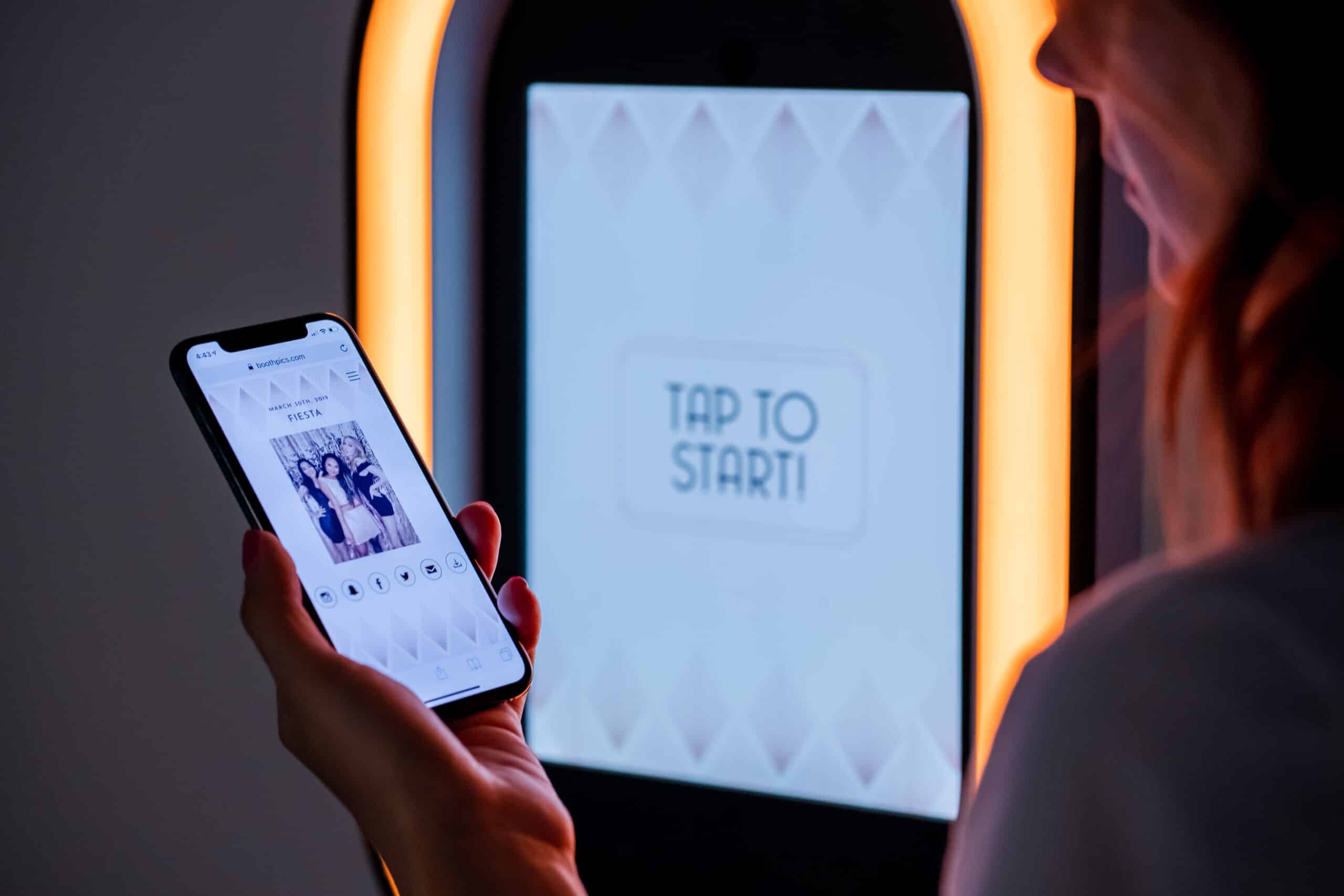 Person holding smartphone displaying photo booth interface with "TAP TO START" prompt, illuminated by soft lighting, emphasizing interactive luxury photo booth experience for events in the DC Metro Area.