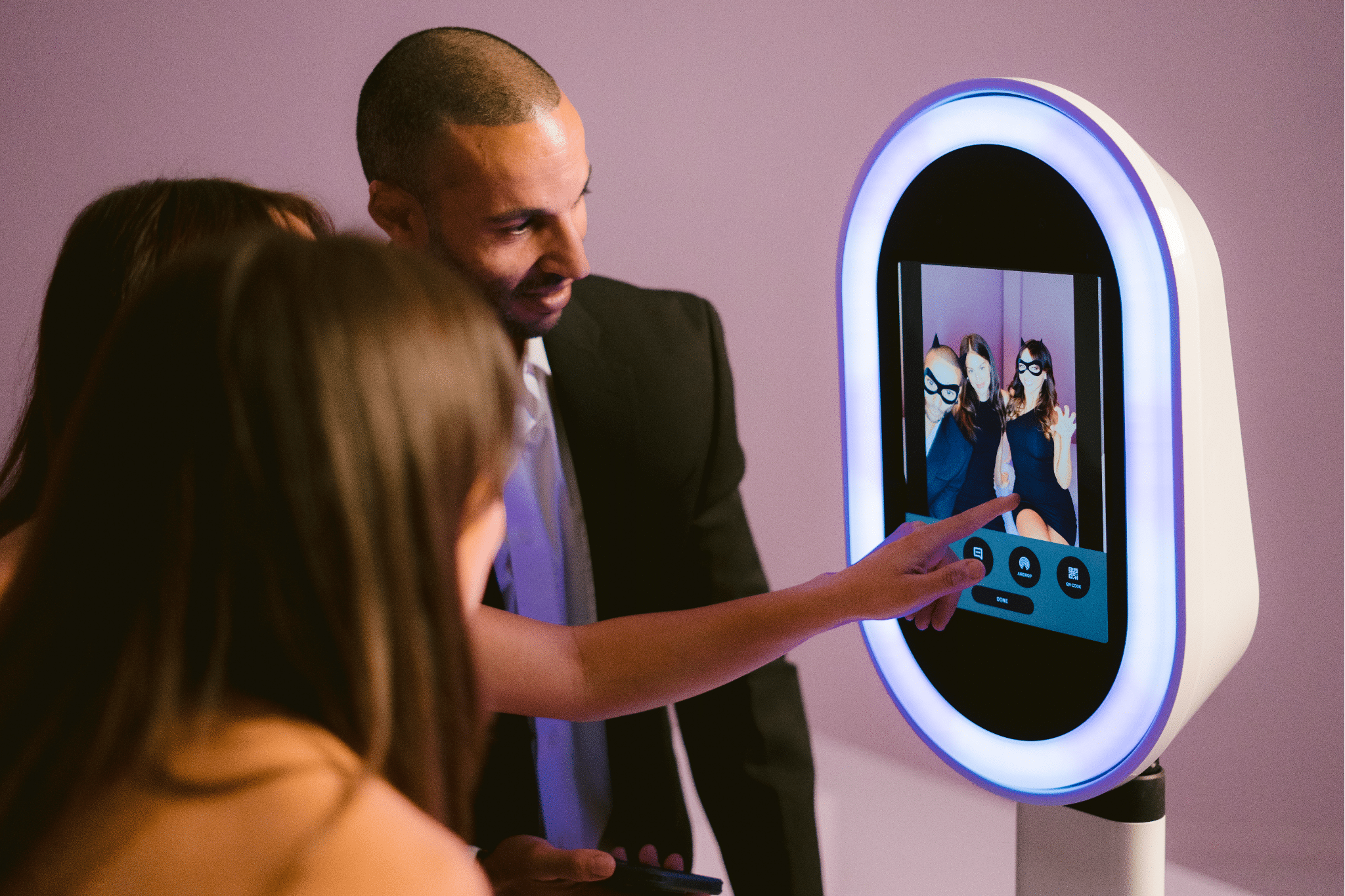 Group of people interacting with a modern photo booth displaying their captured images, featuring customizable options, in a vibrant event setting.