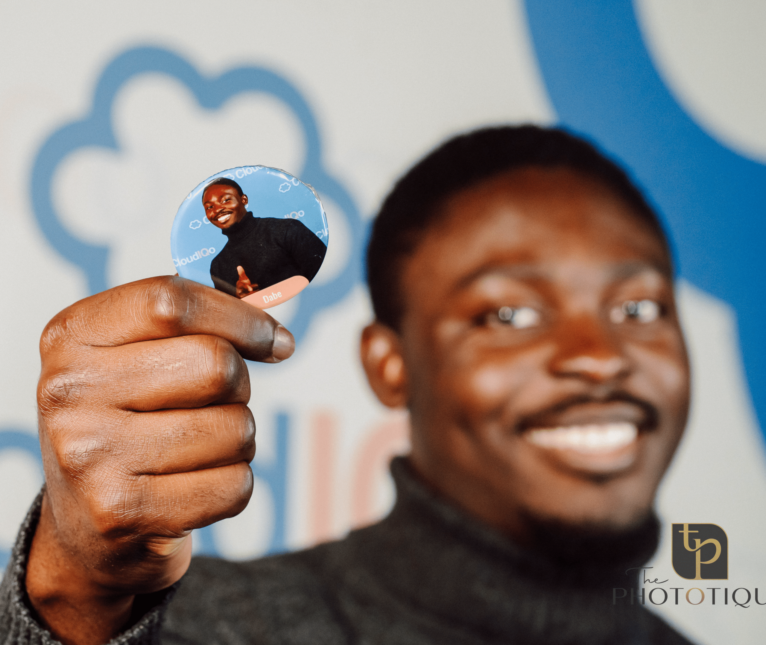 Smiling man holding a branded photo button with a personal image, showcasing engagement at a conference booth, with The Phototique logo in the corner.