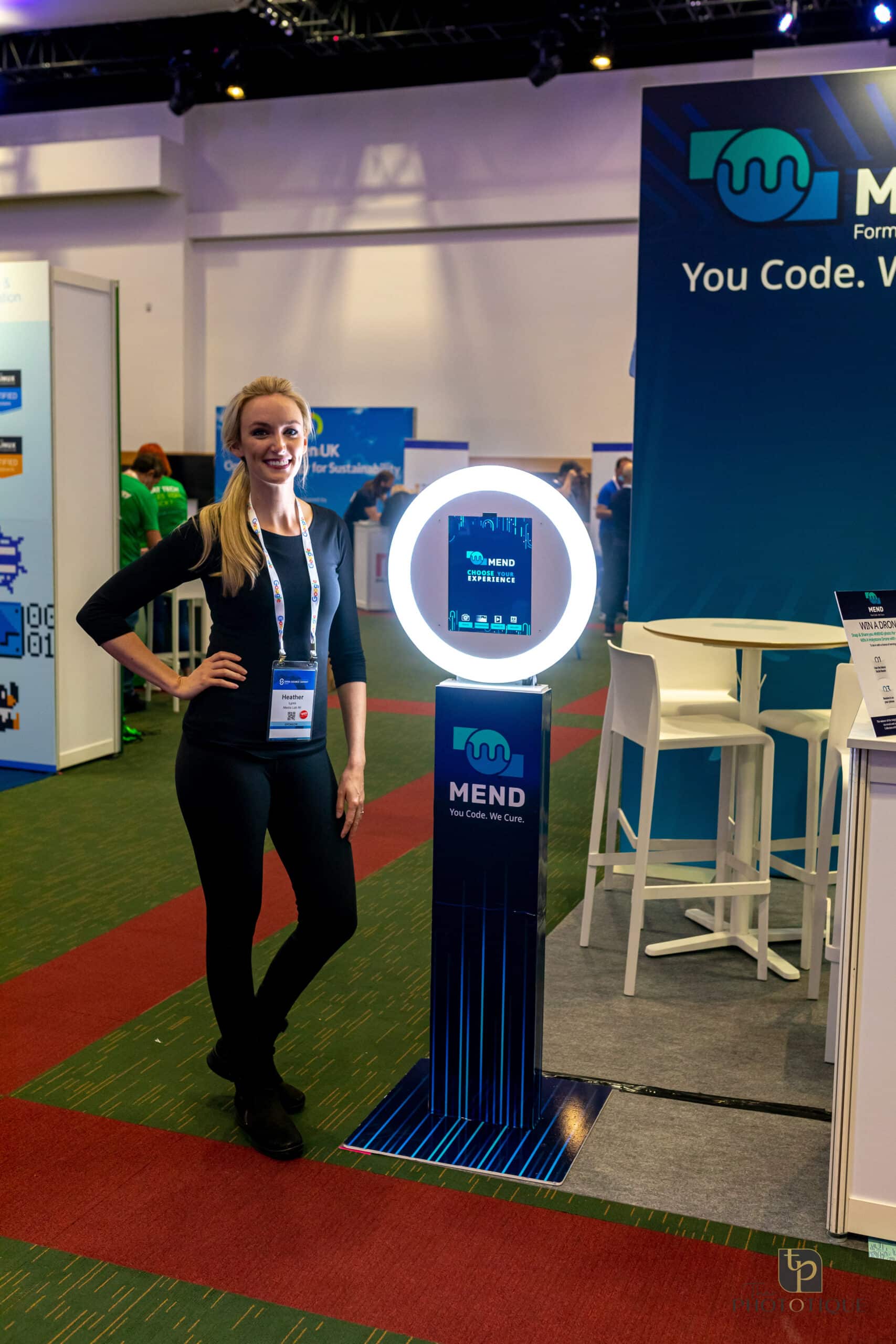 Woman standing next to a photo booth display at a corporate event, showcasing the MEND brand and interactive experience options, with a vibrant exhibition backdrop.