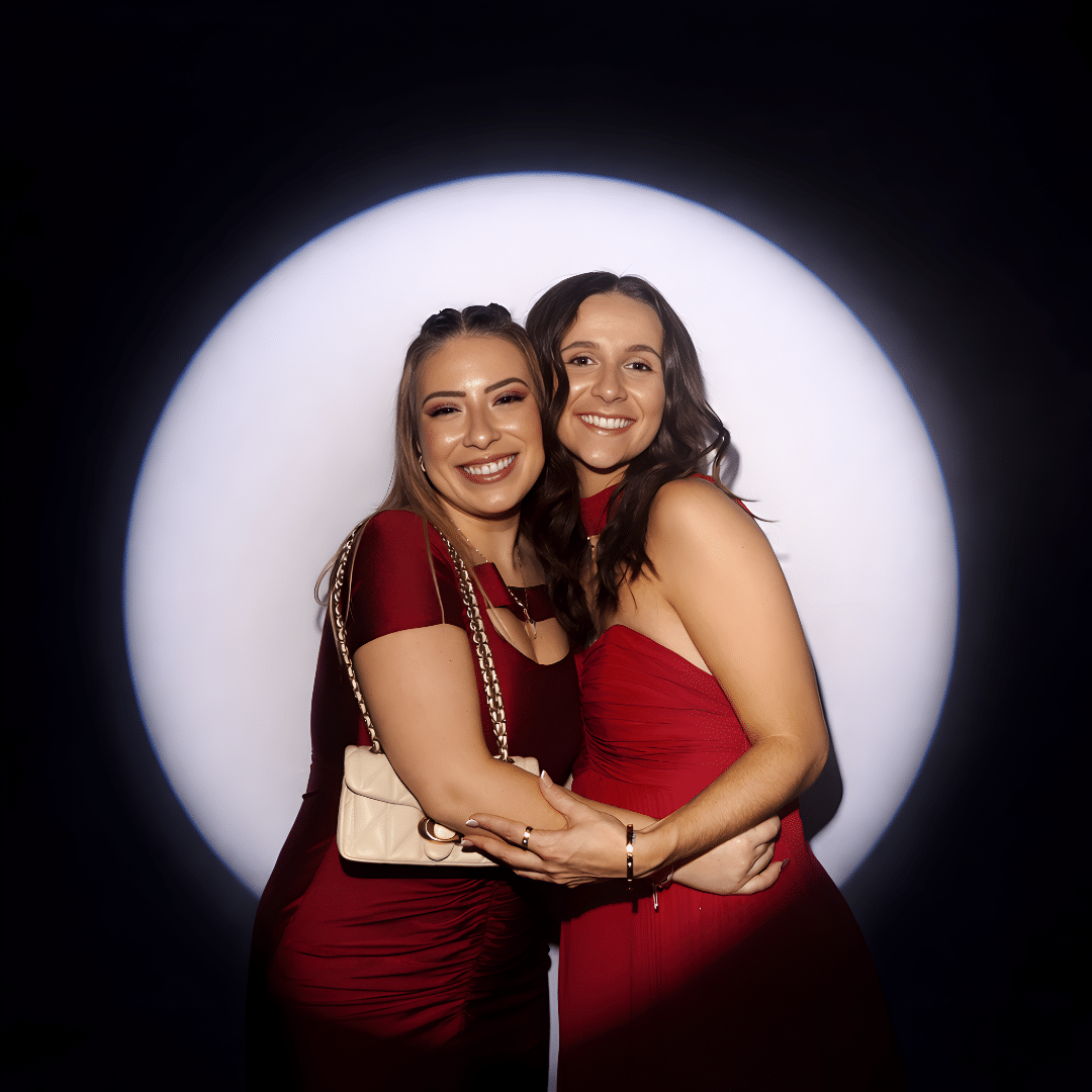 Two women in elegant red dresses smiling and posing together in front of a luxury photo booth backdrop, enhancing the upscale atmosphere of a gala or brand activation event.