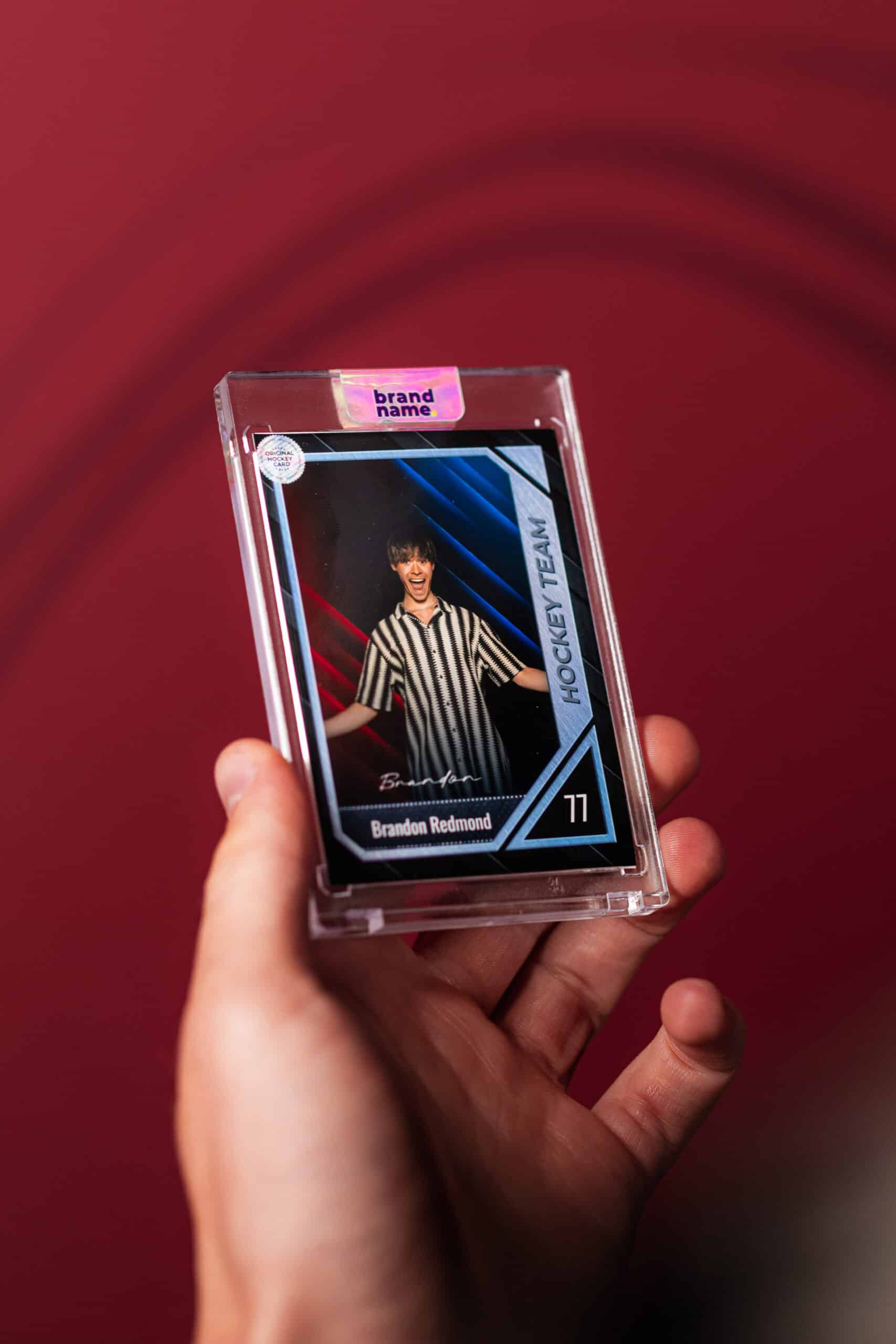 Hand holding a hockey trading card featuring a player in a striped shirt, displaying excitement, with red and blue background elements, emphasizing the photo booth experience offered by The Phototique.